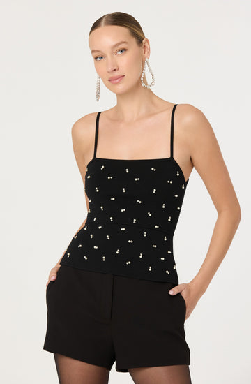 A woman is wearing a black sleeveless top with thin shoulder straps, decorated with small white pearl-like embellishments scattered across the fabric. She pairs it with black shorts and sheer tights. She accessorizes with large, dangling, pearl-encrusted earrings. Her hair is slicked back, and her makeup is natural.