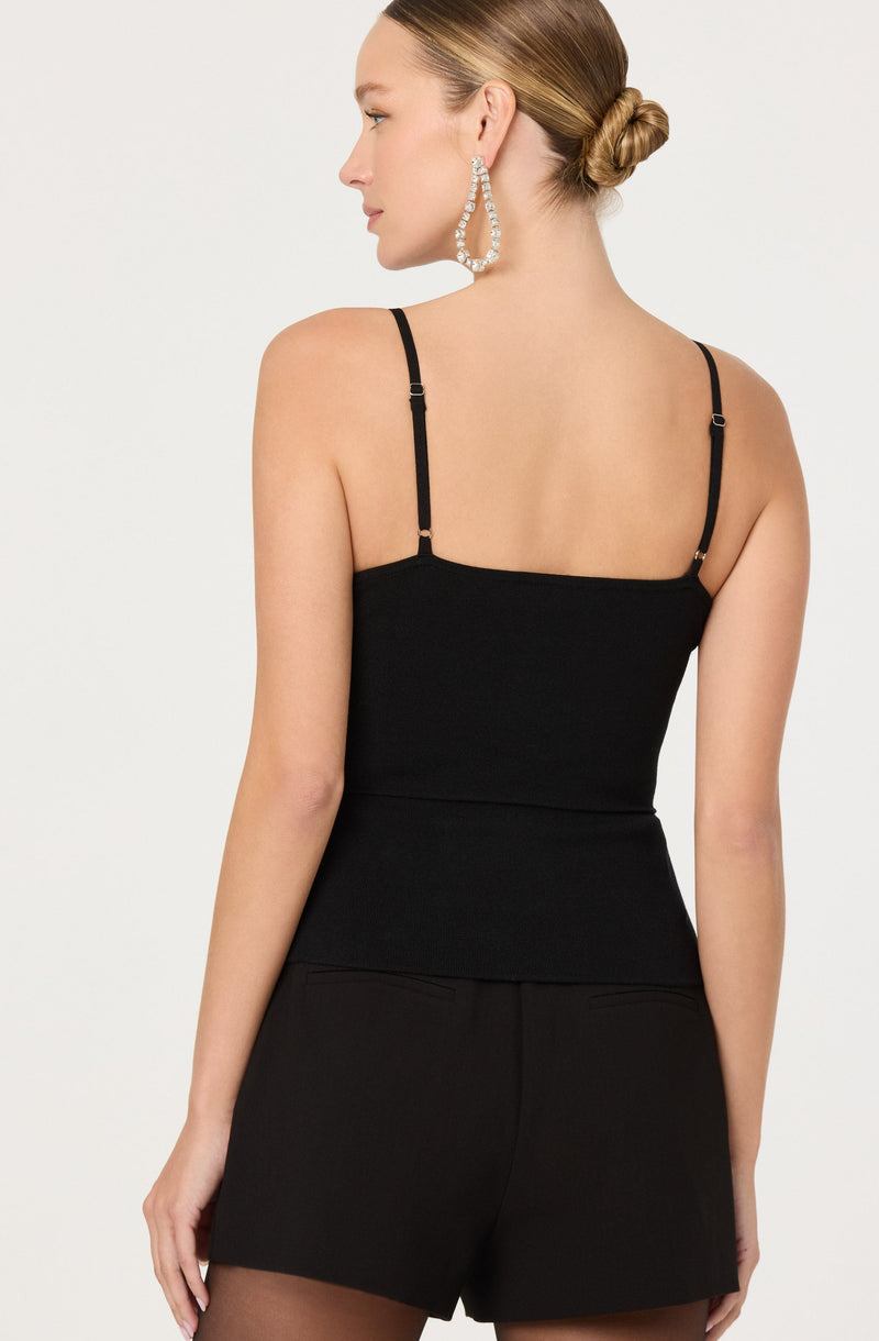 A woman is wearing a fitted black spaghetti strap top and black shorts. She has her hair styled in a low bun and is accessorized with large, statement hoop earrings.