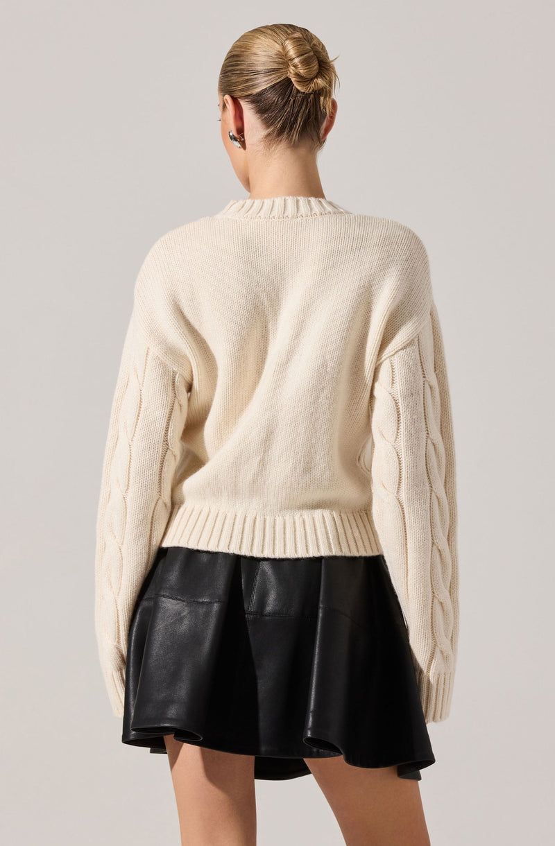 A person is wearing a chunky cream-colored knit sweater with cable knit details running down the sleeves. The sweater has a round neckline and a ribbed hem. They are also wearing a black leather skirt with a flared, A-line shape. The person's hair is styled in a neat bun.