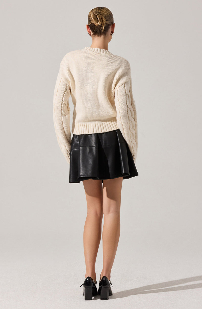 A woman is wearing an oversized, cream-colored knitted sweater with a crew neckline and cable-knit detailing along the sleeves. She pairs it with a high-waisted, black leather skirt that has a flared silhouette. She completes the outfit with black, high-heeled shoes with ankle straps.