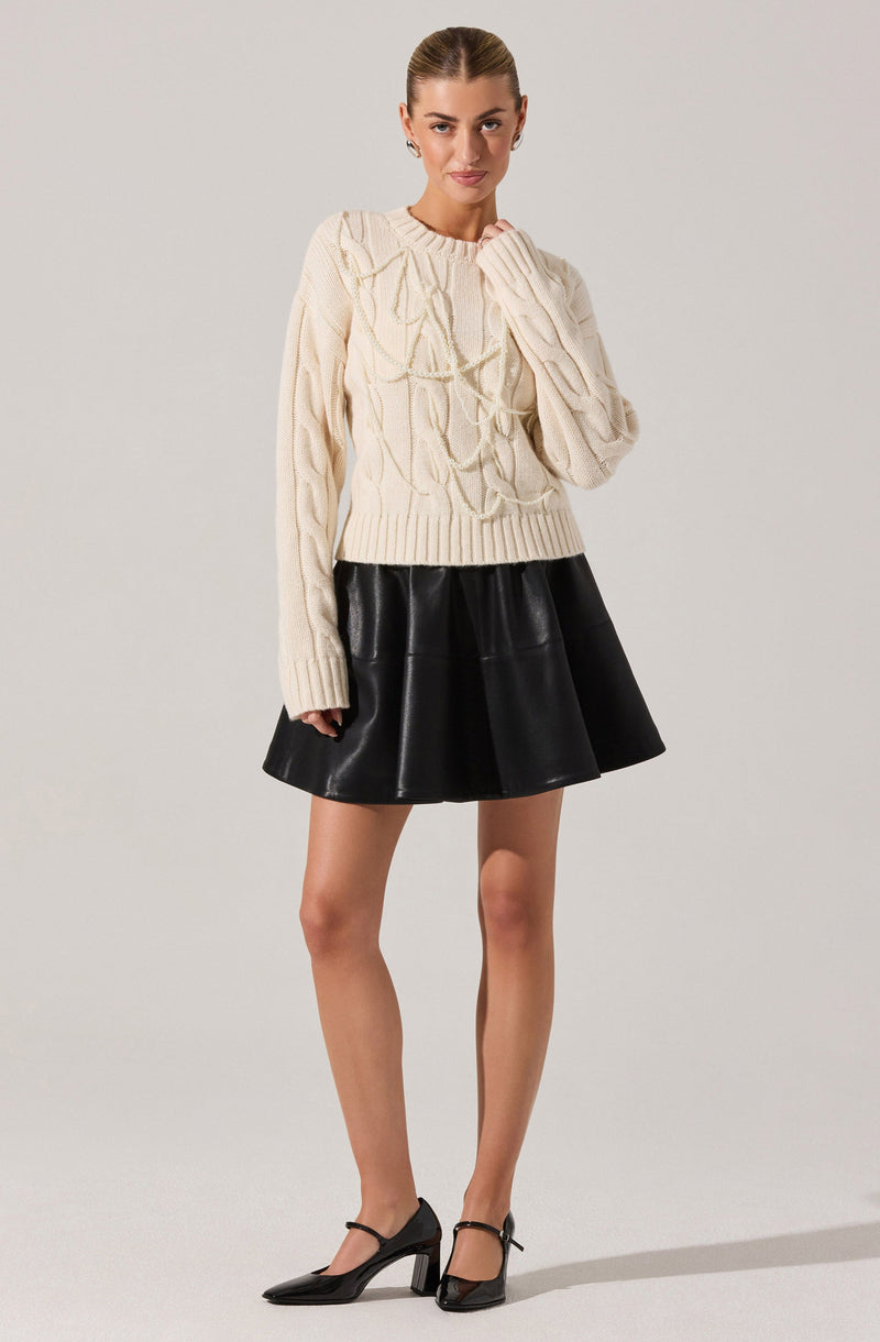 A woman is wearing a cream-colored, chunky knit sweater with a cable pattern and long sleeves. She pairs it with a black, flared, leather-like skirt that falls above her knees. She completes her outfit with black high-heeled shoes featuring ankle straps.
