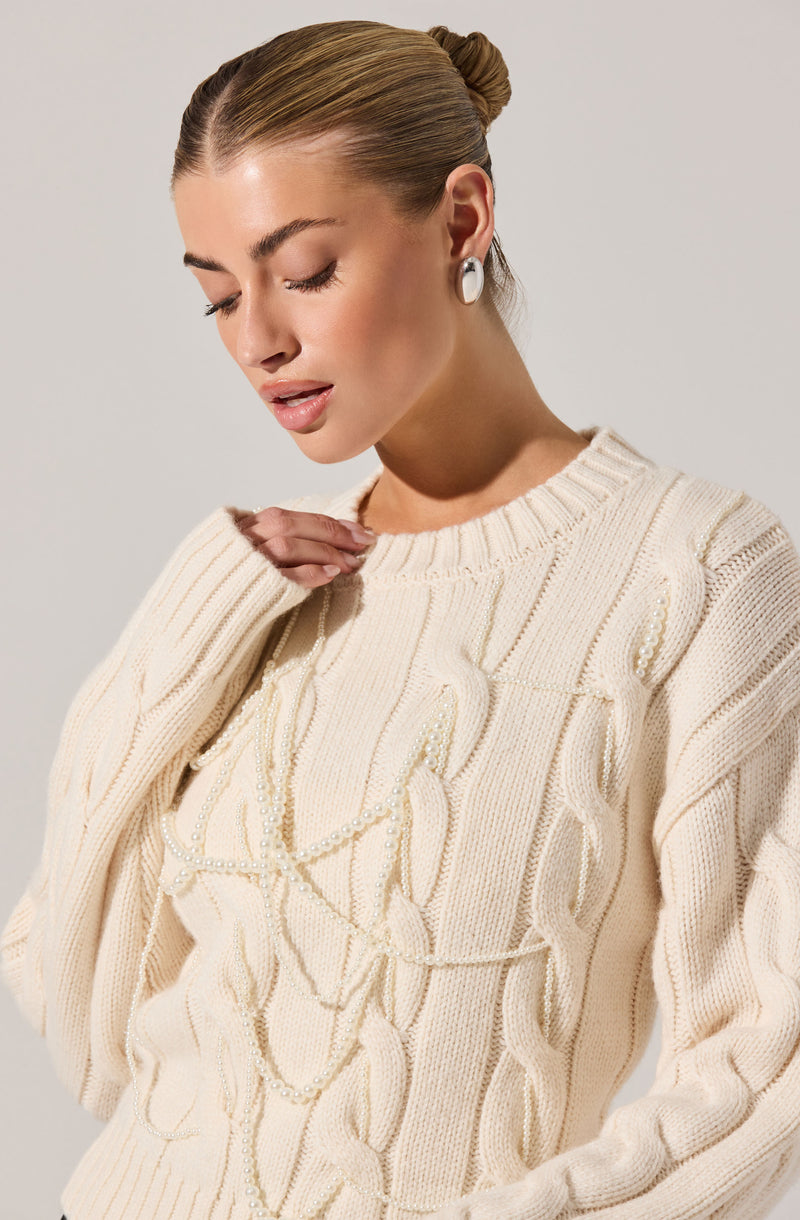 A woman is wearing an oversized cream-colored knitted sweater with a cable-knit pattern and decorative pearl accents. She accessorizes with pearl earrings and her hair is styled in a sleek bun.