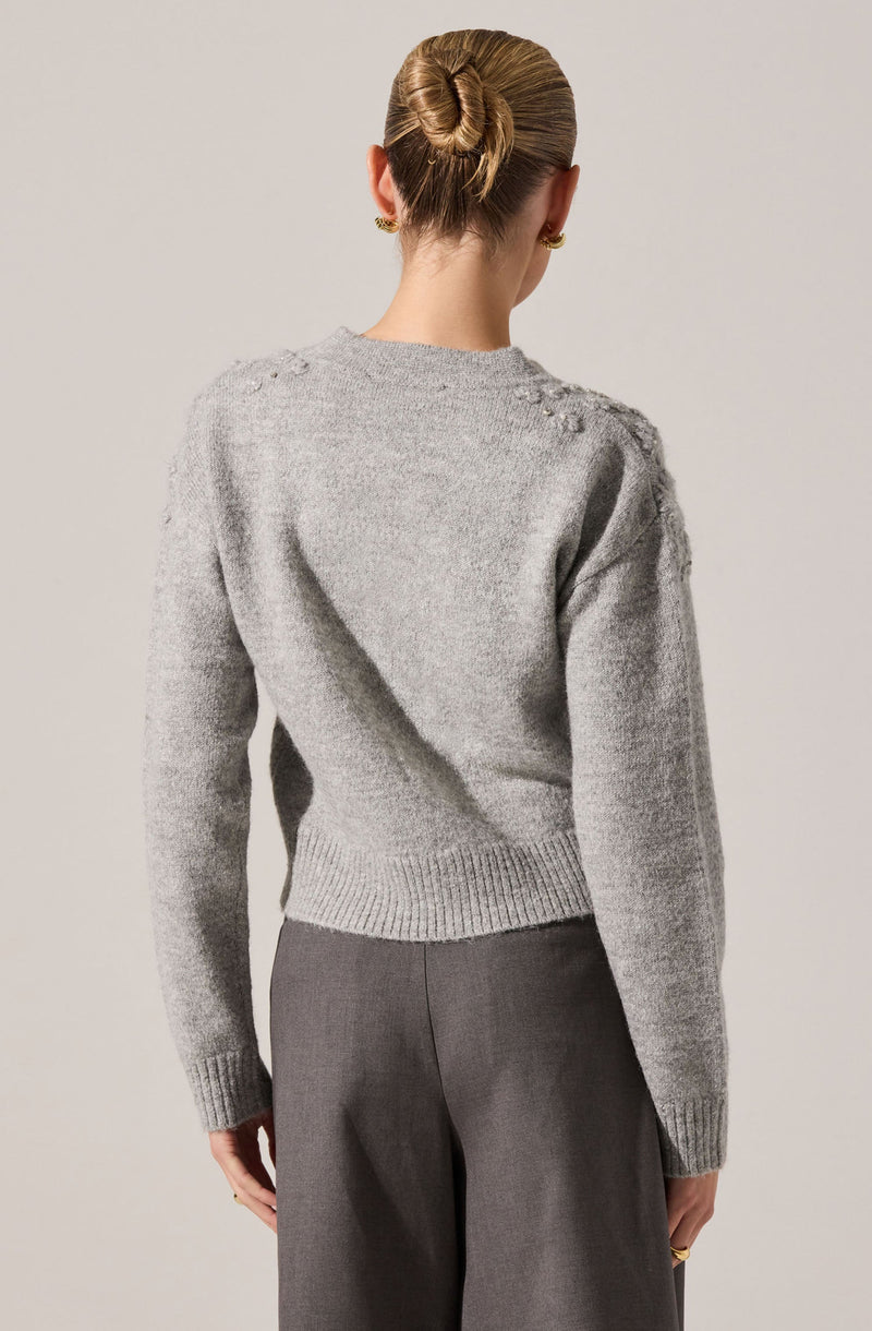 A woman is wearing a light gray, knitted sweater with a crew neckline and ribbed hem and cuffs. The sweater has small, raised decorative details on the shoulders. She is also wearing high-waisted, gray wide-leg trousers. Her hair is styled in a sleek, twisted bun, and she has small gold earrings.
