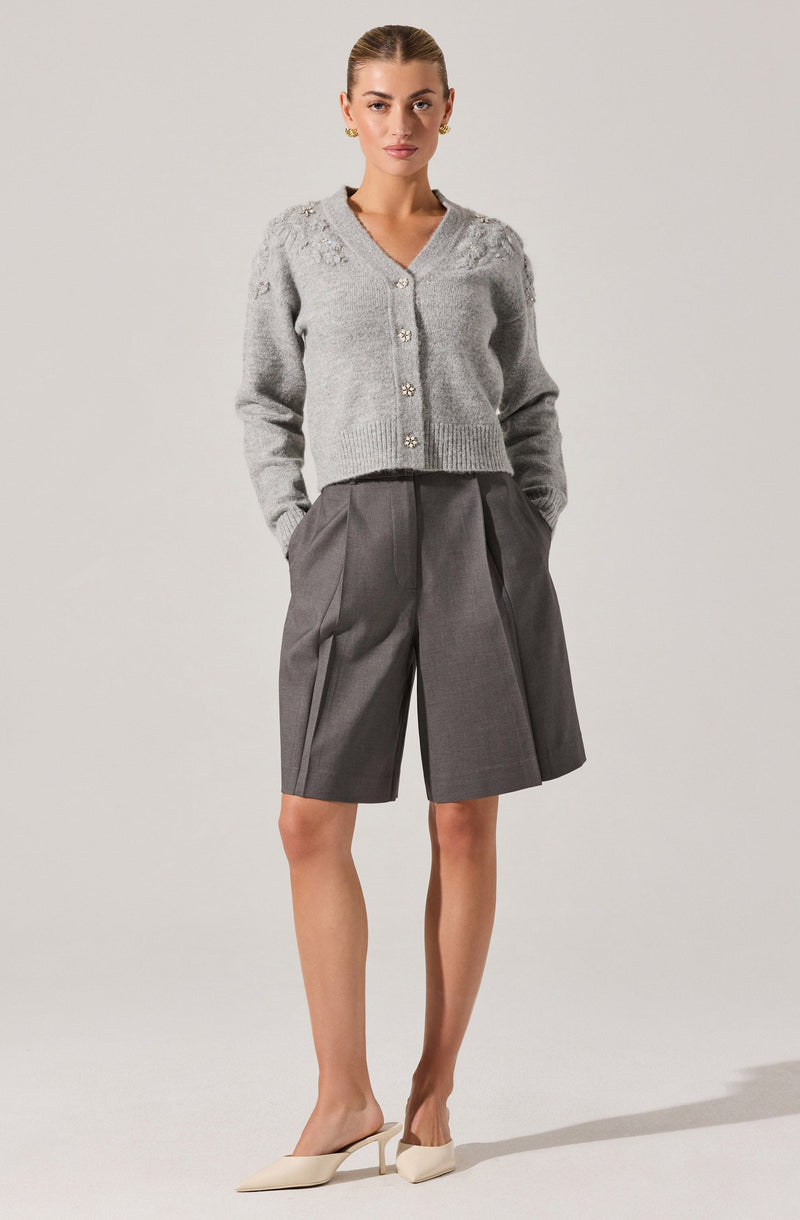 A woman is wearing a light gray, knitted cardigan with a v-neckline and floral embellishments on the shoulders. The cardigan has pearl-like buttons down the front. She is also wearing high-waisted, wide-leg gray shorts with pleats and front pockets. She has on white pointed-toe heels.