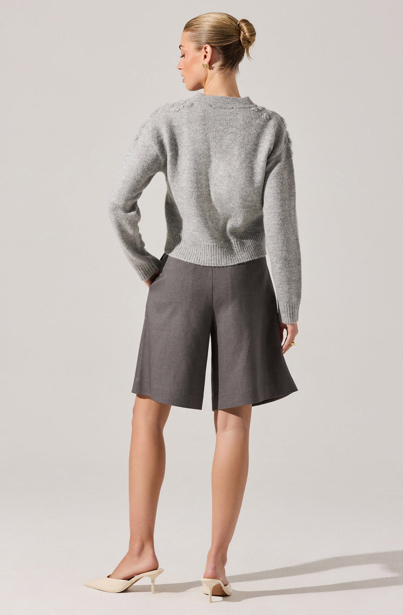 A woman is wearing a light gray, long-sleeved, slightly oversized knit sweater with a ribbed hem and cuffs. She pairs it with high-waisted, wide-leg gray shorts with a subtle texture, featuring a front crease and side pockets. She completes the look with beige pointed-toe high-heeled shoes.