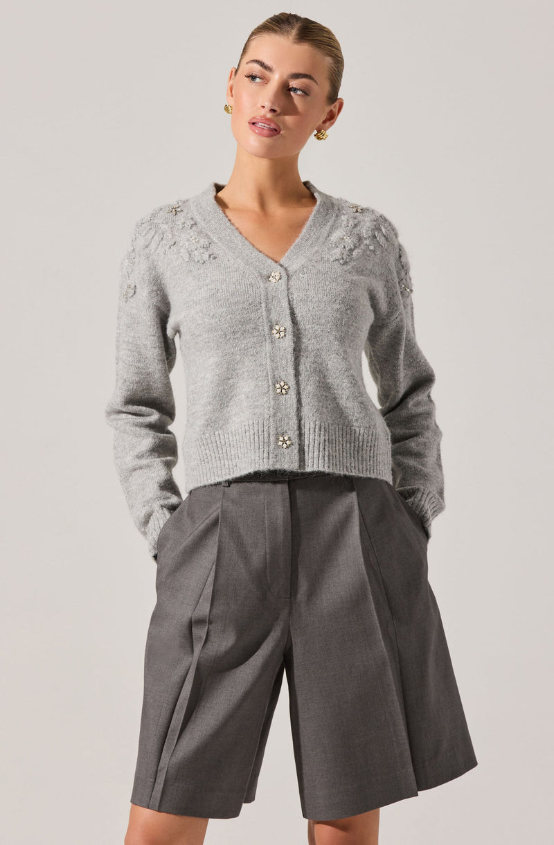 A woman is wearing a light gray, long-sleeved, button-up cardigan with decorative embellishments on the shoulders, paired with high-waisted, knee-length gray shorts that feature pleats and large front pockets.