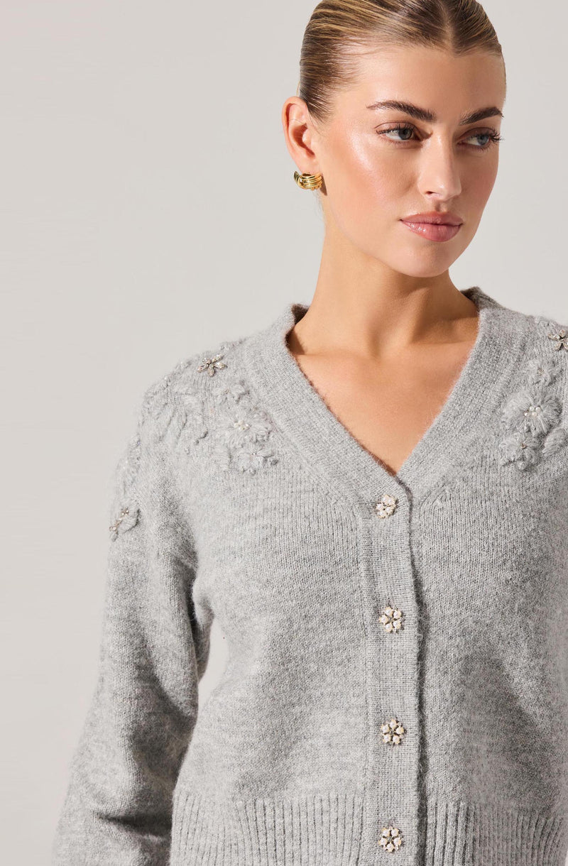 A woman is wearing a light gray knit cardigan with a V-neckline. The cardigan has decorative floral embellishments on the shoulders and down the front, with pearl-like centers and metallic accents.