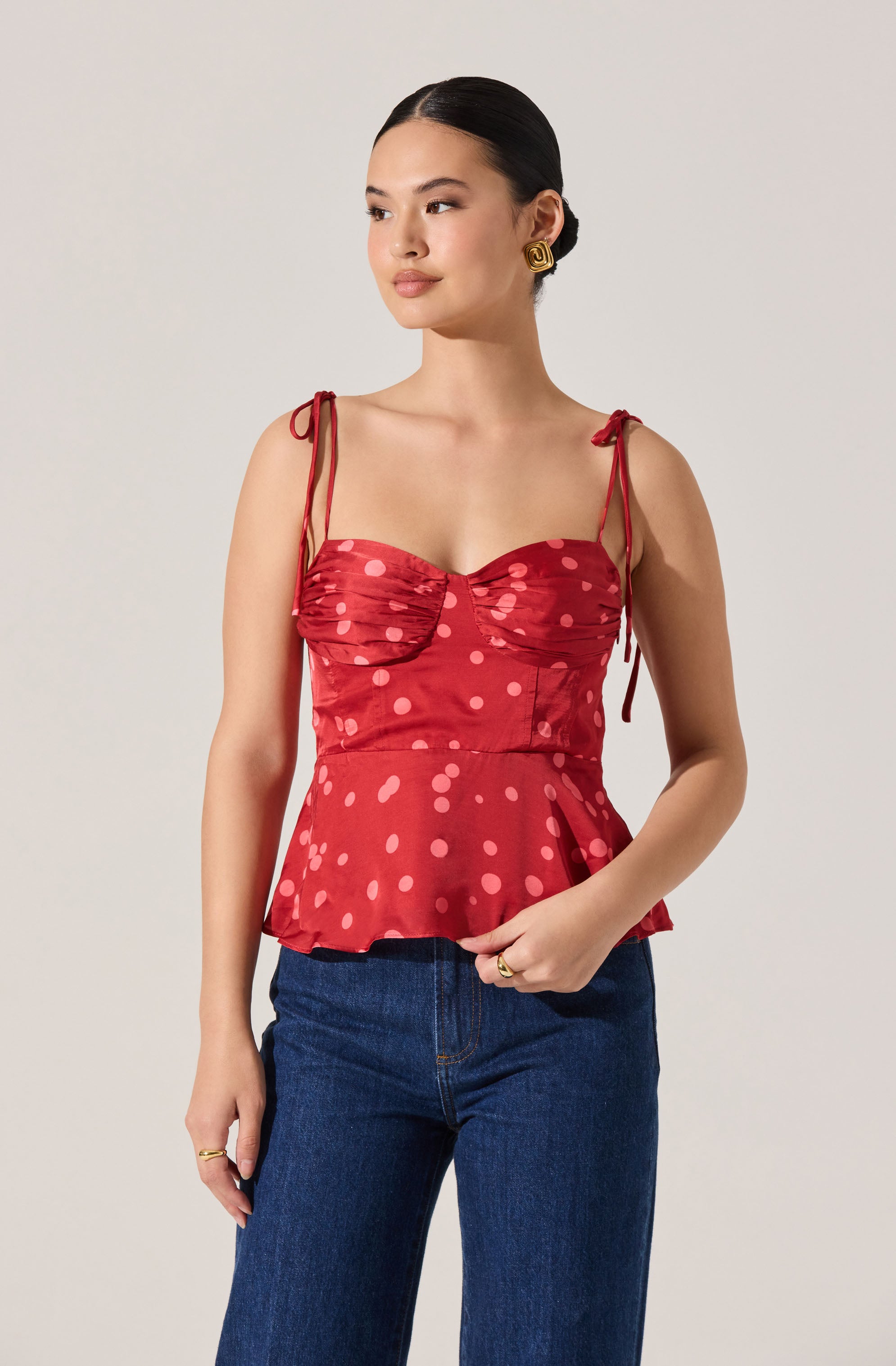 A woman is wearing a sleeveless red top with thin, tied shoulder straps and a sweetheart neckline. The top has a ruched bust area and features a pattern of large pink polka dots. It has a ruffled peplum hem that flares out at the waist. She is also wearing dark blue jeans.