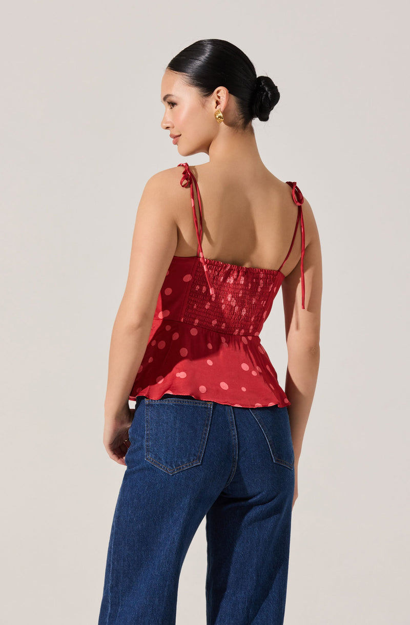 A woman is wearing a sleeveless red top with thin, tied shoulder straps. The top features a smocked bodice and a ruffled peplum hem with a polka dot pattern. She is also wearing high-waisted, dark blue jeans.