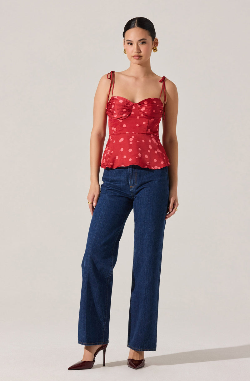 A woman is wearing a red sleeveless top with thin shoulder straps tied into bows, a sweetheart neckline, and a peplum hem. The top features a pattern of pinkish dots. She pairs it with high-waisted, straight-leg blue jeans and pointed brown high heels.
