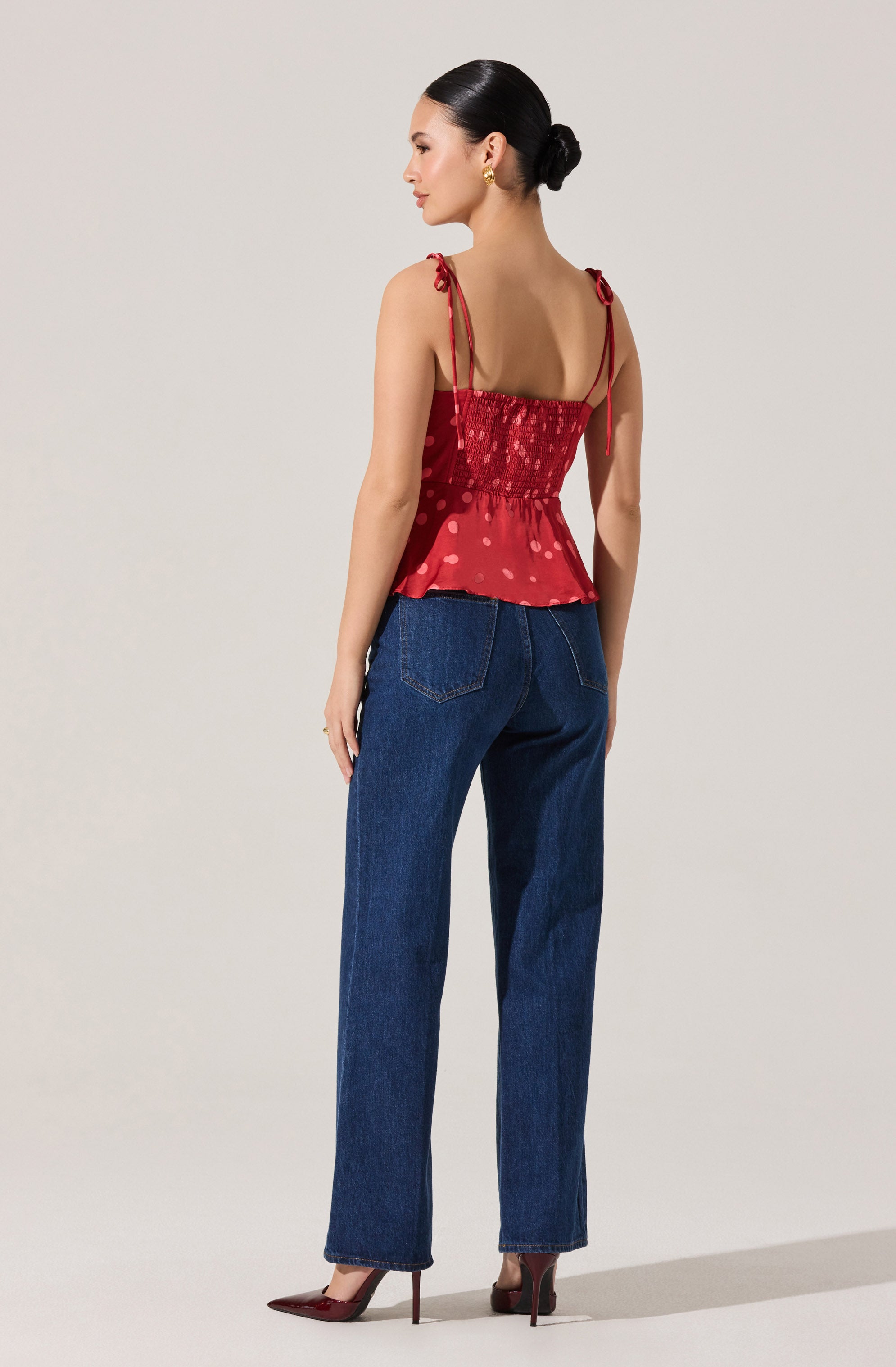 A woman is wearing a red sleeveless top with adjustable shoulder straps tied in bows, featuring a smocked bodice and a peplum hem with a subtle polka dot pattern. She pairs it with high-waisted, straight-leg dark blue jeans and matching red high heels.