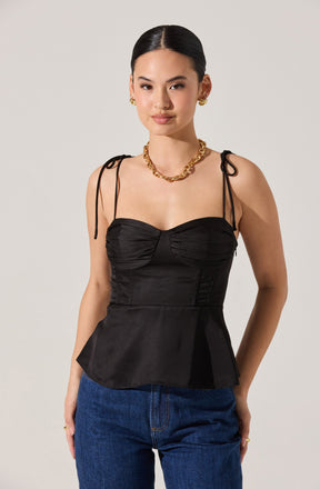 A woman is wearing a fitted black satin top with ruched bust detailing and thin, adjustable shoulder ties. The top has a peplum waist and is paired with blue jeans. She accessorizes with gold jewelry, including a chunky chain necklace, matching earrings, and rings.