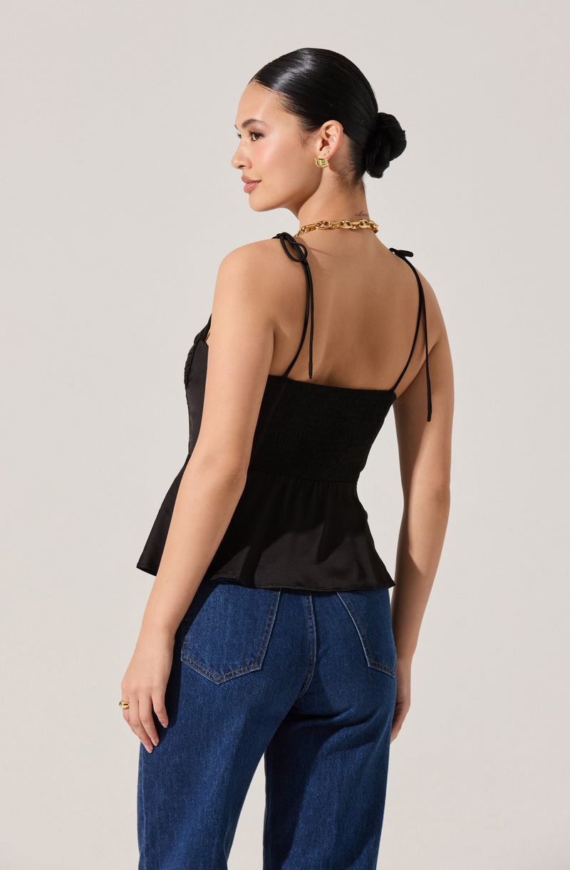 A woman is wearing a black spaghetti strap top with tie straps on the shoulders, featuring a fitted, gathered waist and a peplum hem. She pairs the top with high-waisted blue jeans.