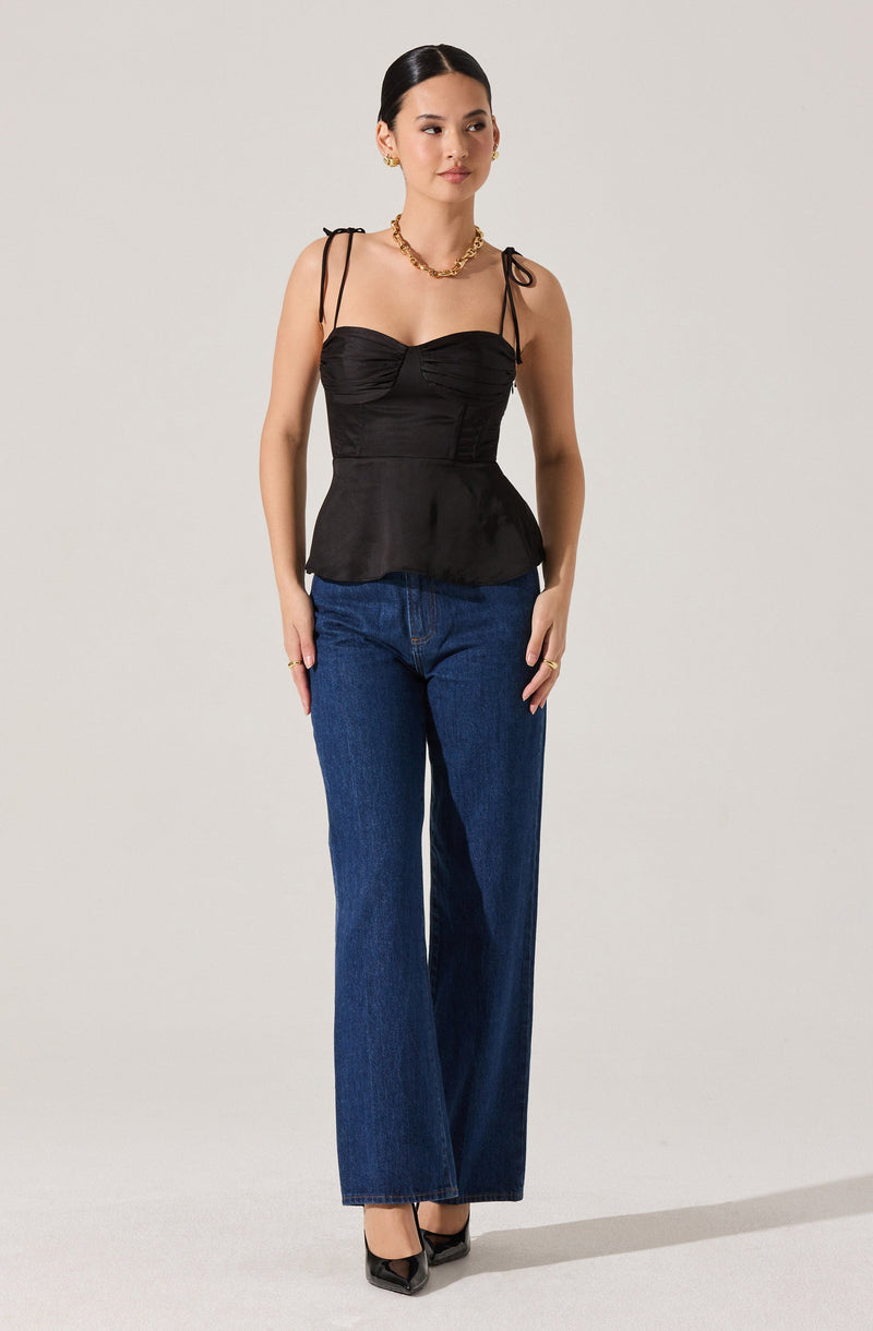 A woman is wearing a black, fitted, sleeveless top with ruched detailing on the bust and thin, tied shoulder straps. She pairs it with high-waisted, wide-leg blue jeans and black pointed-toe high heels. She accessorizes with gold jewelry, including earrings, a necklace, and rings.
