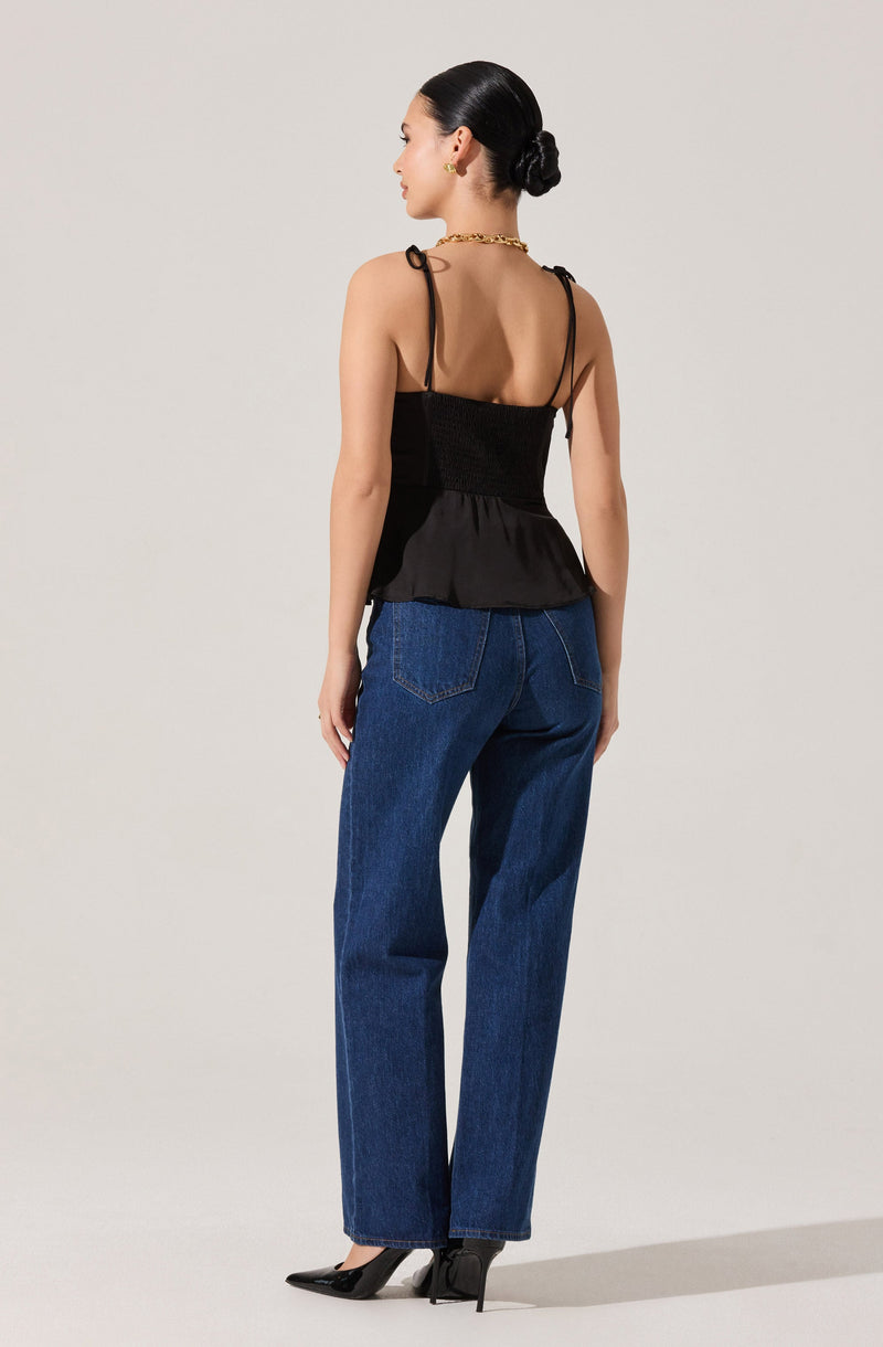 A woman is wearing a black sleeveless top with thin spaghetti straps and a peplum hem. The top features a textured, smocked bodice and a flowy lower section. She pairs it with high-waisted, straight-leg blue jeans. She is also wearing black pointed-toe high heels. Her hair is styled in a sleek, low bun, and she accessorizes with a gold chain necklace and earrings.