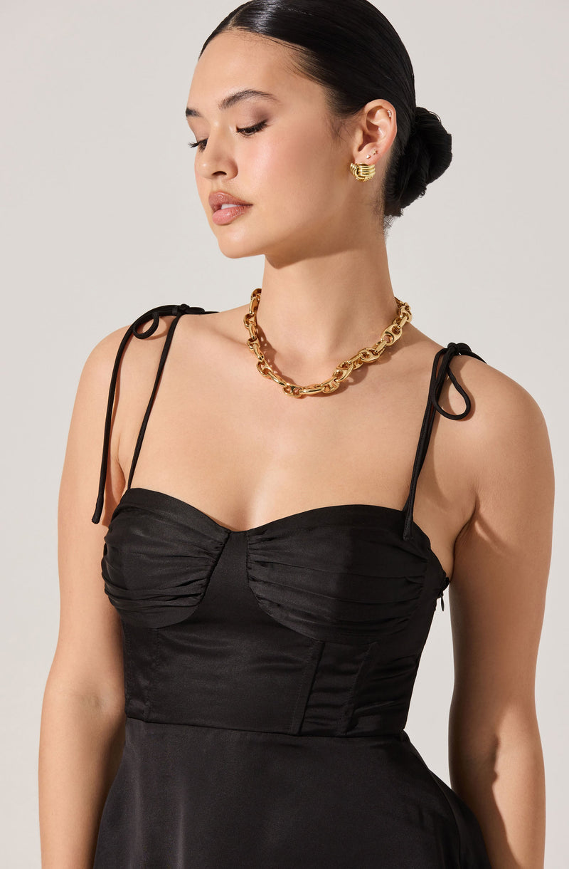 A woman is wearing a black dress with ruched detailing on the bust area and thin, adjustable shoulder ties. She accessorizes with a chunky gold chain necklace, gold earrings, and has her dark hair styled in a sleek, low bun.
