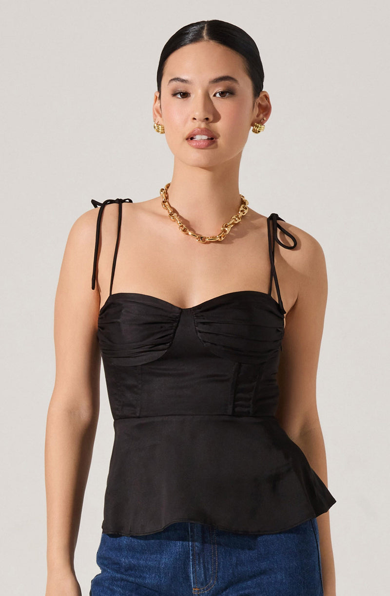 A woman is wearing a black sleeveless top with ruched detailing across the bust area, thin shoulder straps tied into bows, and a peplum hem. She has paired it with blue jeans and accessorized with a chunky gold chain necklace and gold hoop earrings.