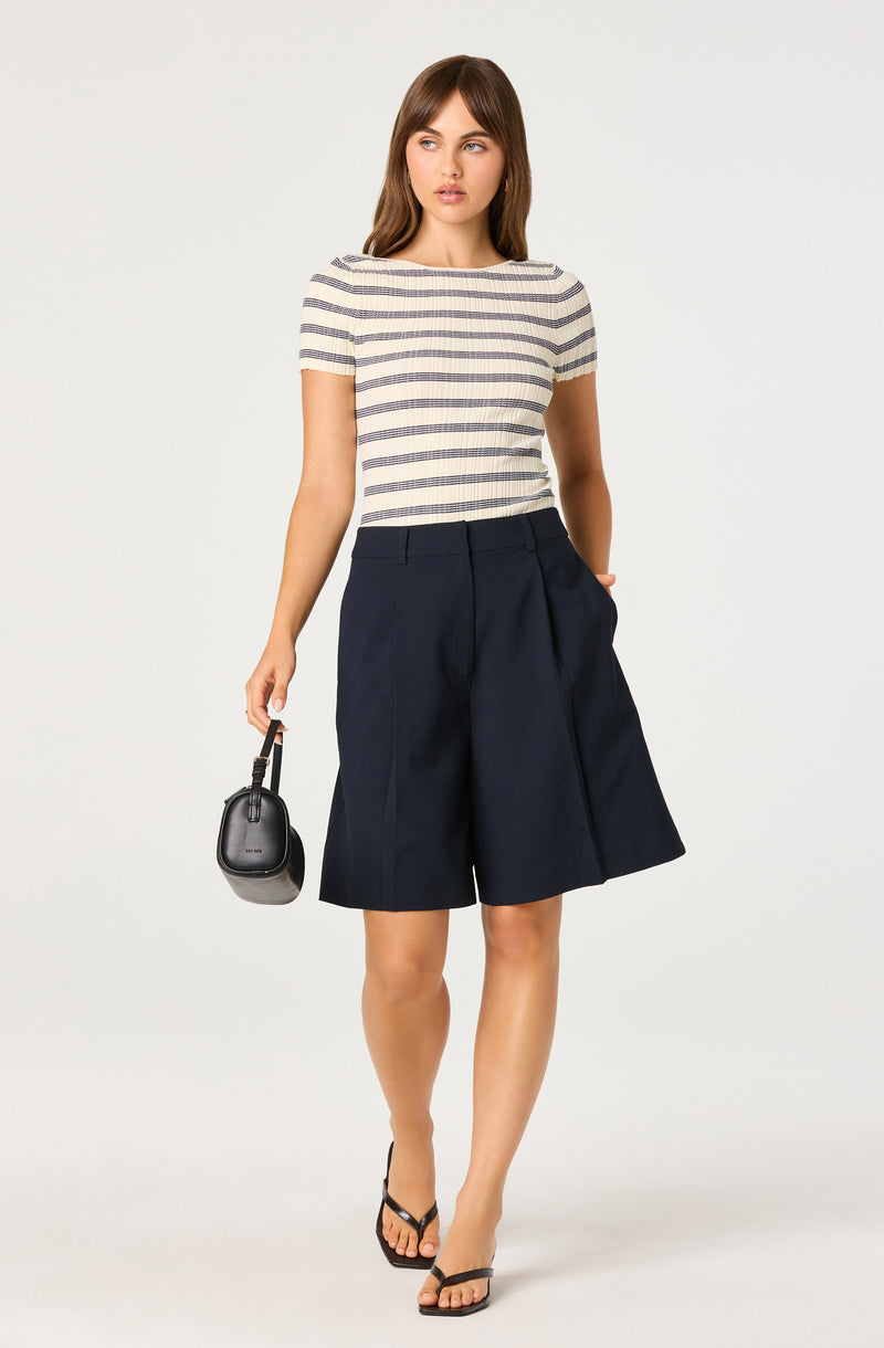 Woman in striped top and navy shorts holding a small black handbag, wearing black sandals.