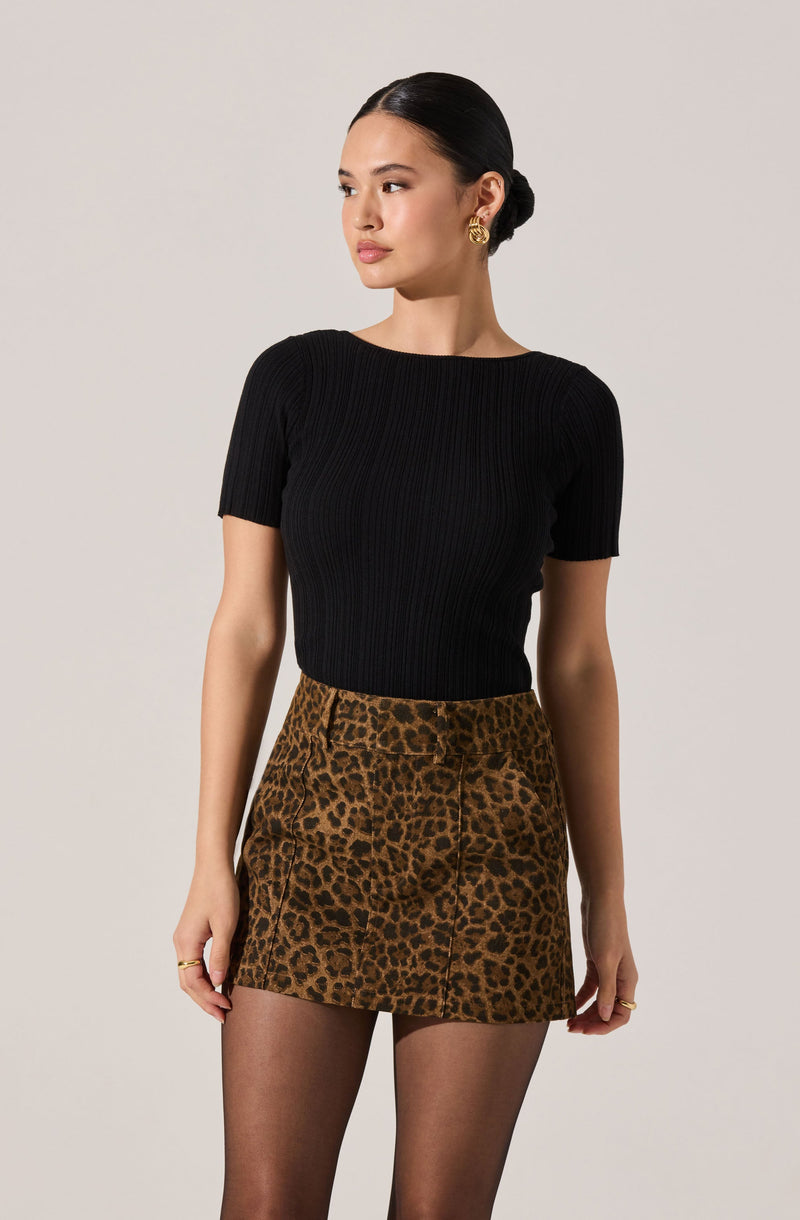 A woman is wearing a fitted black ribbed short-sleeve top tucked into a high-waisted leopard print mini skirt. She also has on black tights and gold jewelry, including earrings and rings.