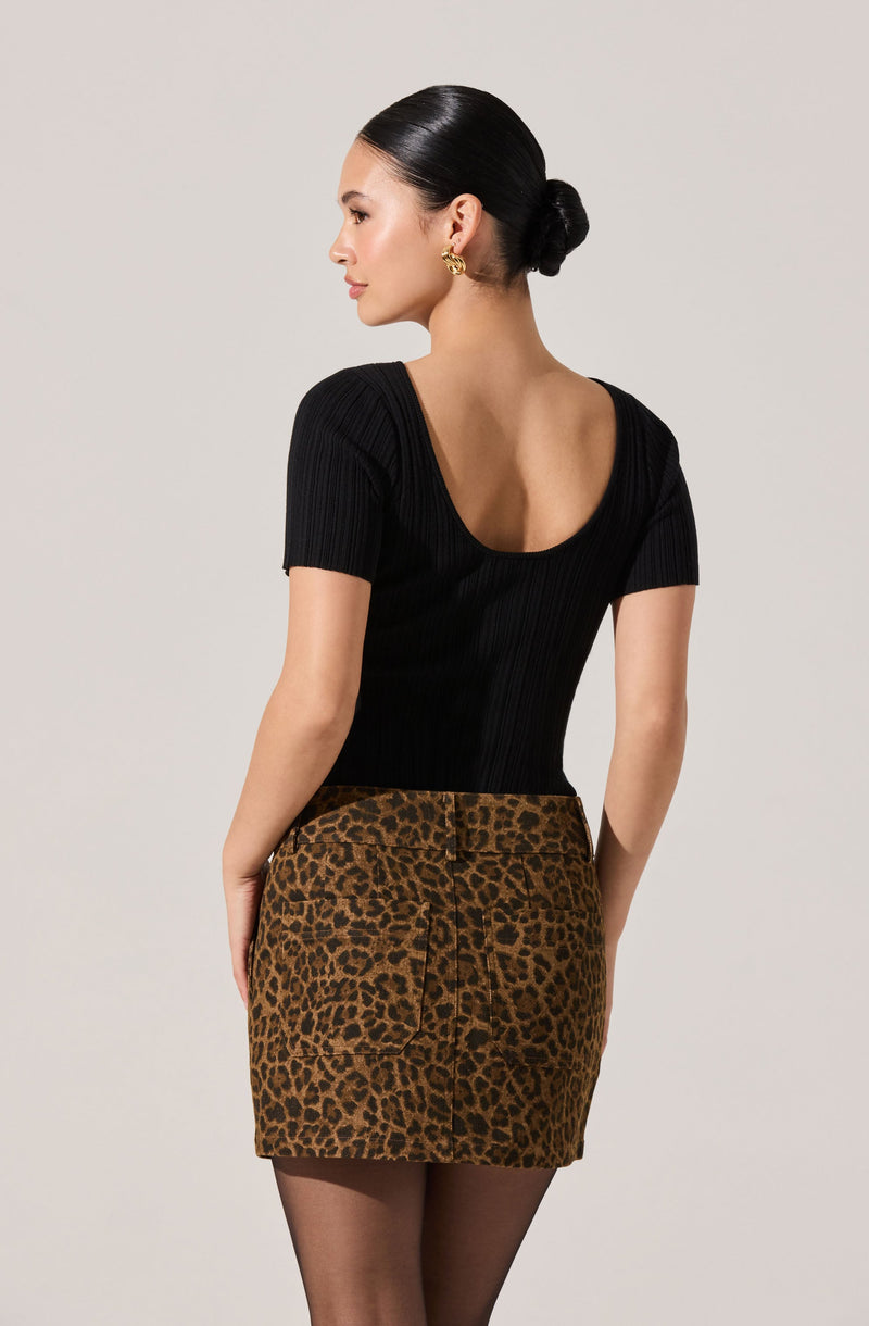 A woman is wearing a fitted black short-sleeve top with a deep scoop back and ribbed texture, paired with a high-waisted leopard print skirt with front pockets.