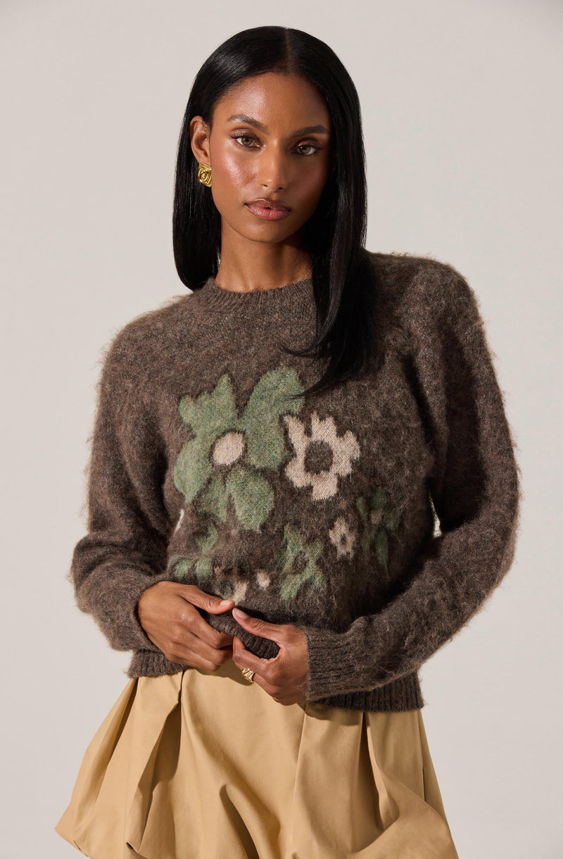 A woman is wearing a dark brown fuzzy sweater with a floral pattern in green, cream, and black. The sweater has a round neckline and long sleeves. She pairs it with a beige pleated skirt. She has straight black hair and is accessorized with gold earrings.