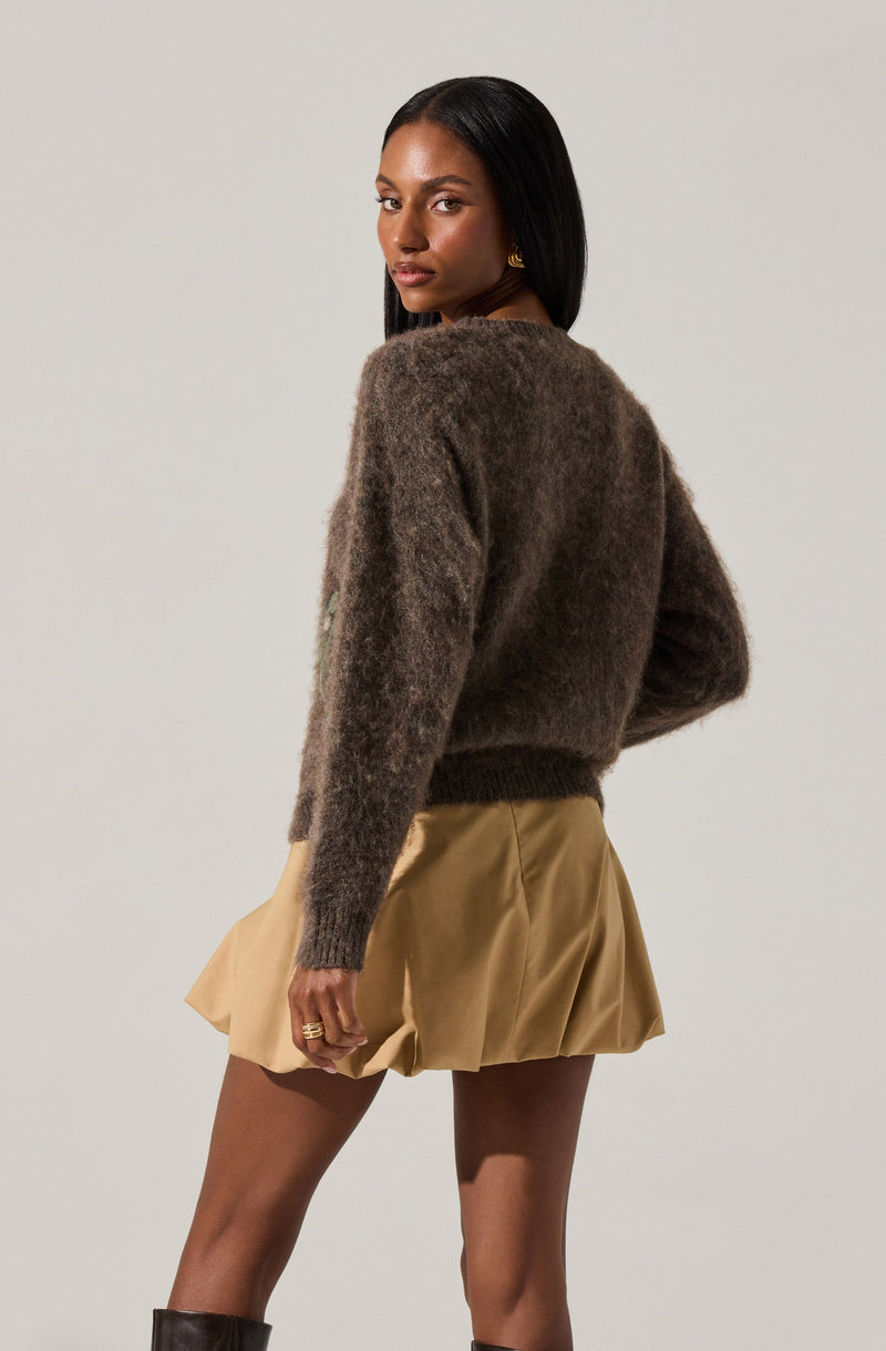 A woman is wearing a loose, long-sleeved, fuzzy, dark brown sweater and a high-waisted, beige, pleated mini skirt.