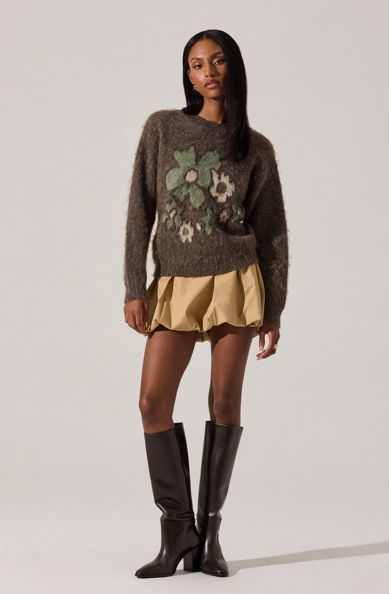 A woman is wearing a dark brown fuzzy sweater with a large floral pattern in light green, white, and beige on the front. She has on a beige, puffy, knee-length skirt with gathered fabric, and black knee-high boots with a pointed toe and a block heel.