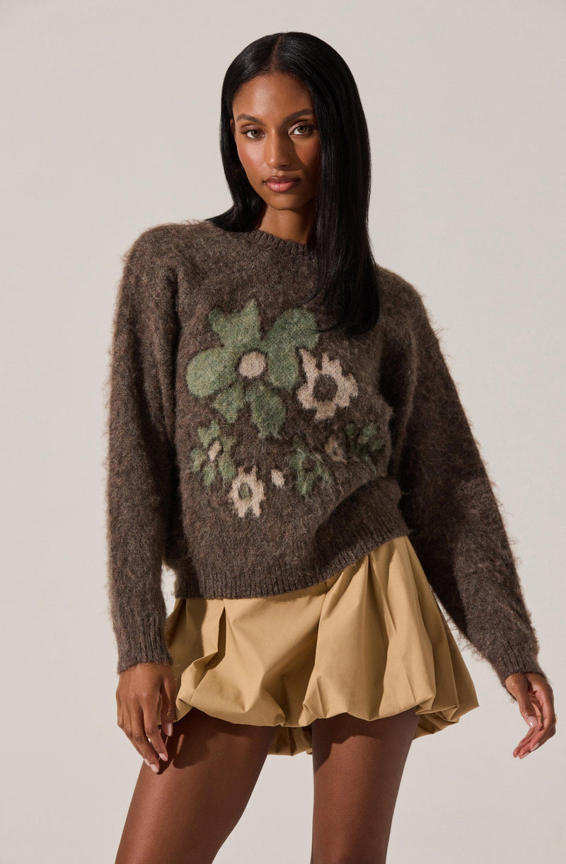A woman is wearing a cozy, oversized brown sweater with large, abstract floral patterns in shades of green, white, and beige on the front. She pairs it with beige, voluminous, bubble-style shorts.