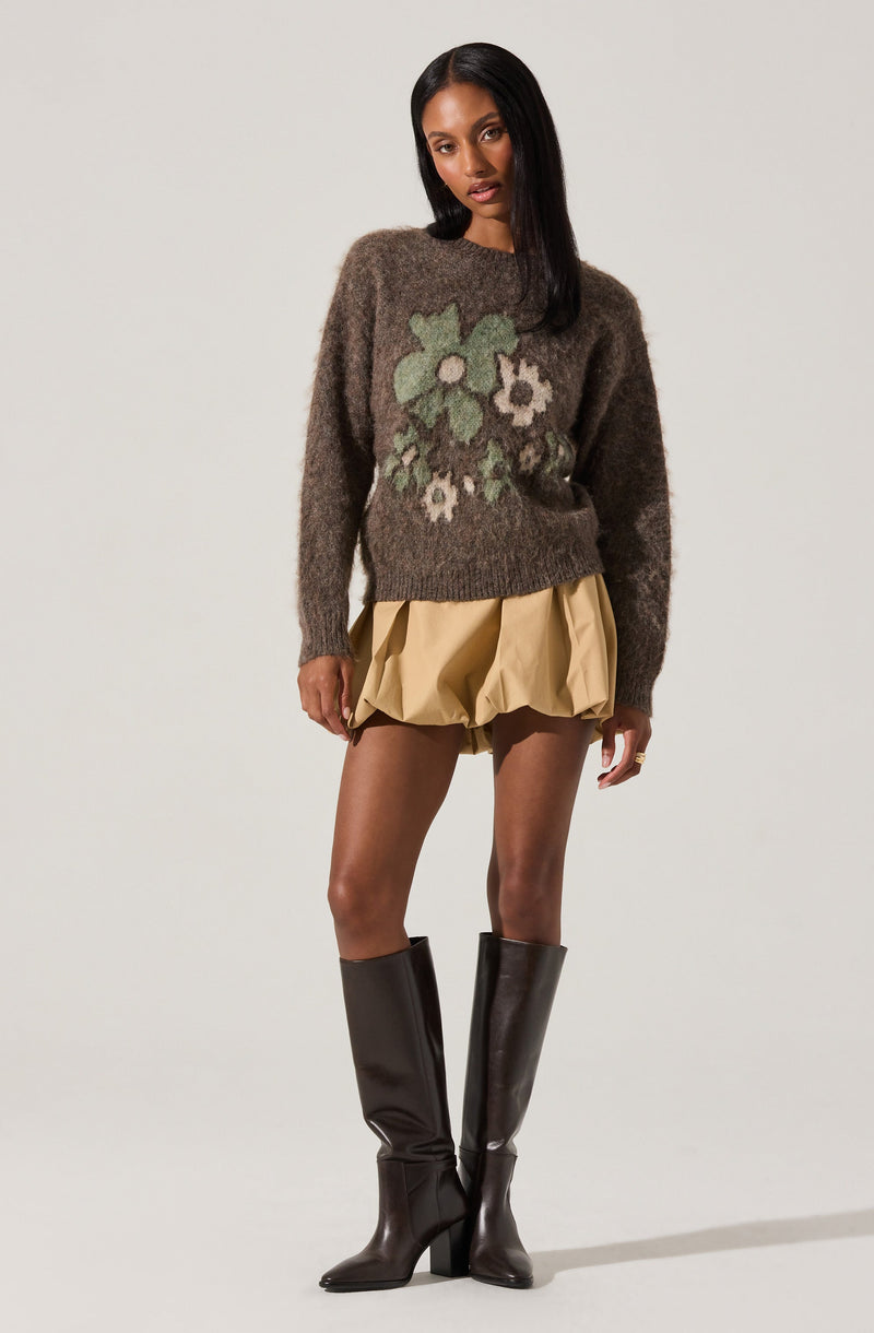 A woman is wearing a fuzzy, oversized brown sweater with a large floral pattern in shades of green, white, and gray on the front. She pairs it with beige, puffy, bubble-style shorts and tall black leather boots with block heels.