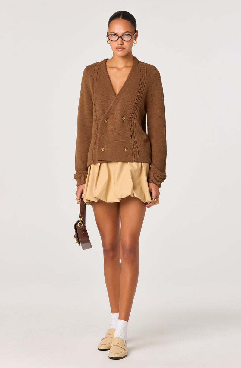 A woman is wearing a brown, double-breasted knit cardigan with gold buttons, featuring a V-neckline and long sleeves. She pairs it with a beige, ruffled, above-the-knee skirt. She accessorizes with glasses, gold hoop earrings, a small brown handbag, white socks, and beige shoes.