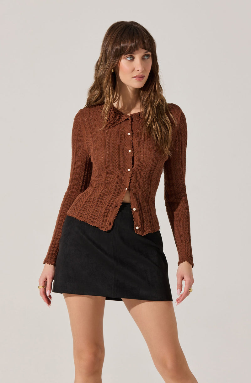 A woman is wearing a fitted, long-sleeved, brown cable-knit cardigan with a collar and pearl buttons down the front. She pairs it with a black A-line mini skirt.
