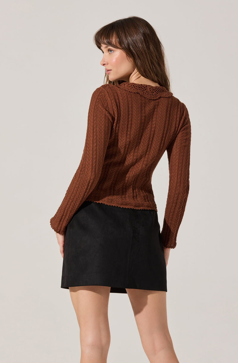 A woman is wearing a brown, long-sleeved, cable-knit sweater with a decorative collar. She pairs it with a black, high-waisted, mini skirt.