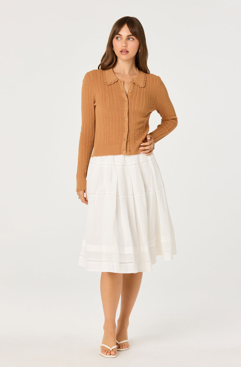 Woman wearing a light brown cardigan with buttons and a white midi skirt, beige sandals.