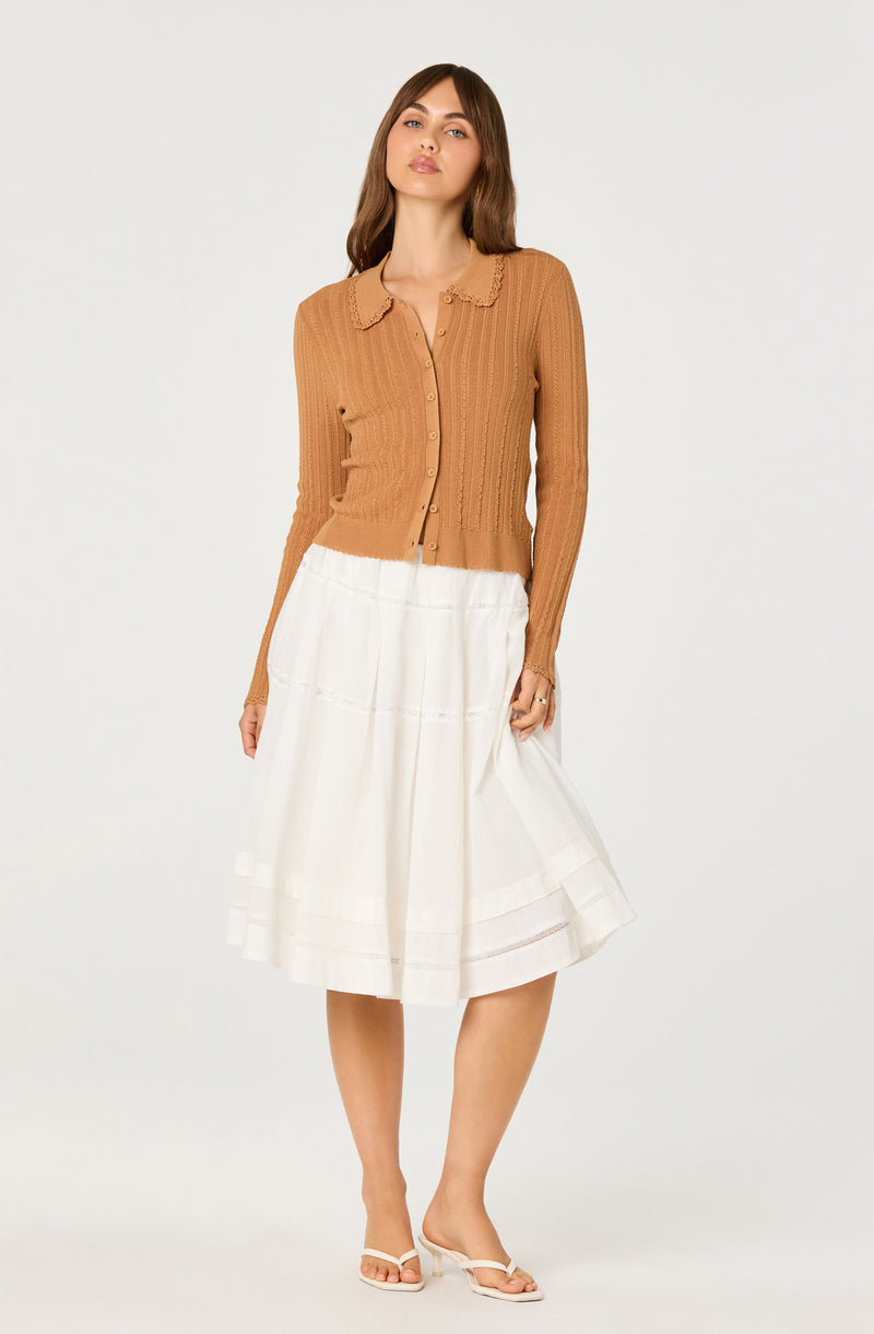 Woman wearing a tan ribbed button-up cardigan with a lace-collared neckline and a white knee-length pleated skirt.