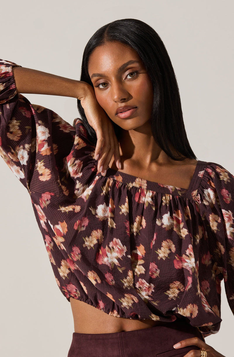 A woman is wearing a dark brown, long-sleeved blouse with a floral pattern in shades of pink, beige, and orange. The blouse has a gathered, square neckline and a loose fit, with the hem ending at her waist.