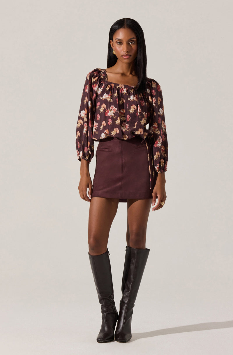 A woman is wearing a long-sleeved, loose-fitting blouse with a floral pattern in shades of pink, cream, and brown on a dark background. The blouse has a square neckline and slightly puffed sleeves. She pairs it with a high-waisted, burgundy mini skirt made of a smooth fabric, featuring a side zipper and a decorative chain detail hanging from the waist. She completes her outfit with tall, black leather knee-high boots.