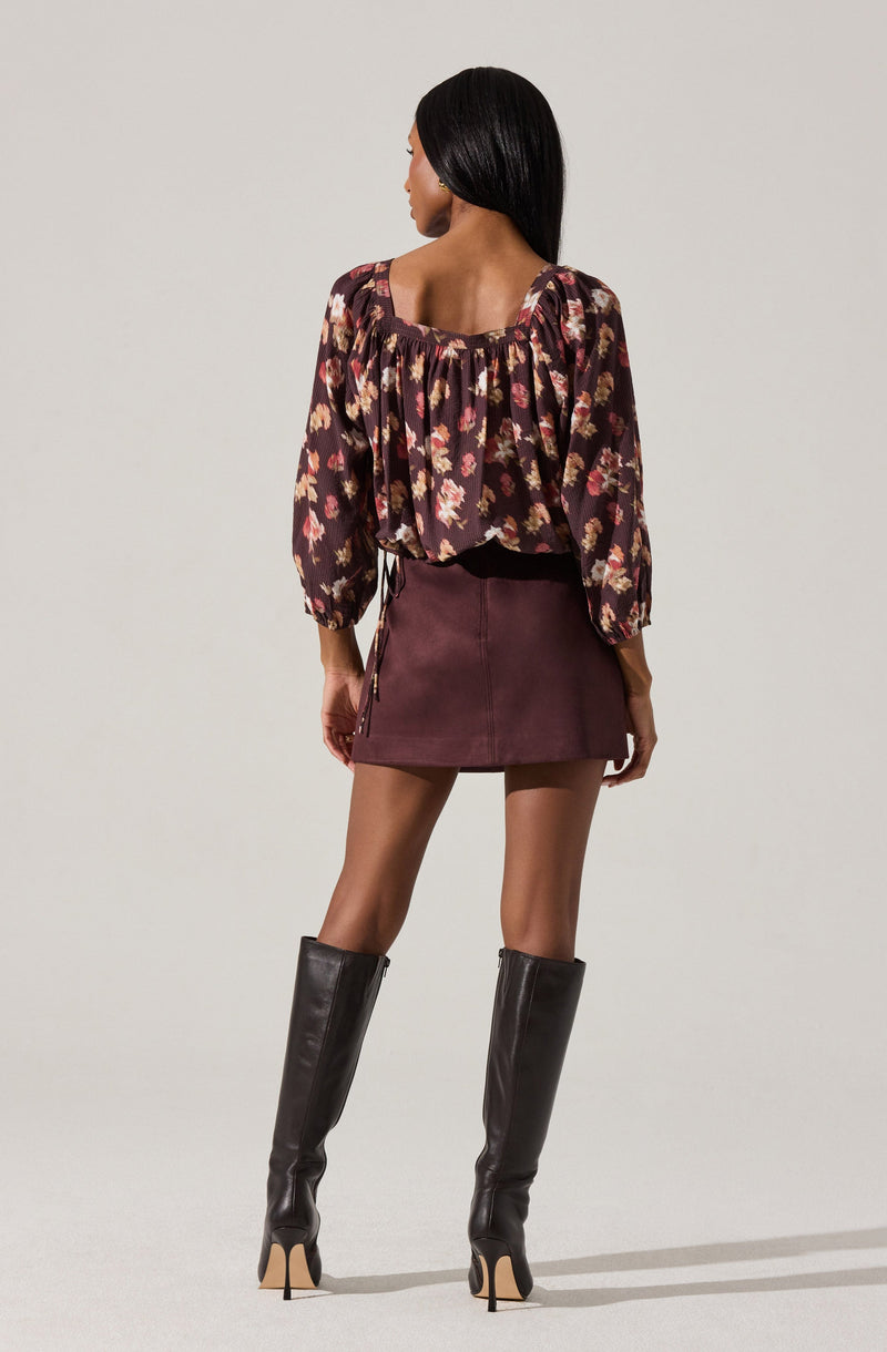 A woman is wearing a long-sleeved, floral-patterned blouse with a square neckline. She pairs it with a high-waisted, burgundy mini skirt and tall black leather boots with high heels.