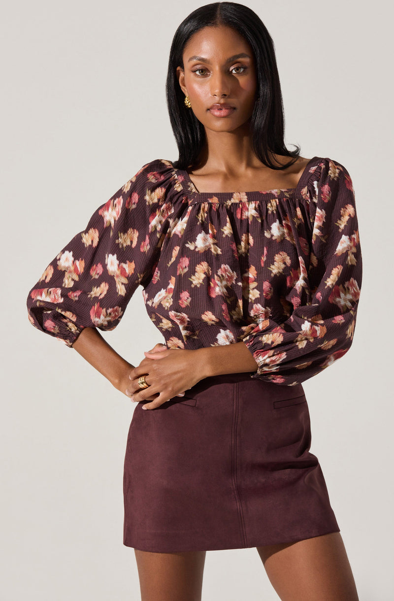 A woman wearing a dark floral print blouse with puffed, three-quarter length sleeves and a square neckline. She pairs it with a high-waisted, burgundy suede mini skirt.