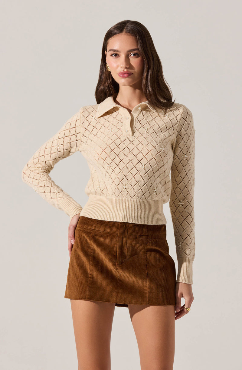 A woman is wearing a cream-colored knit sweater with a diamond pattern and a collar. The sweater has a zip-up neckline and ribbed cuffs and hem. She is also wearing a high-waisted mini skirt made of brown corduroy fabric.