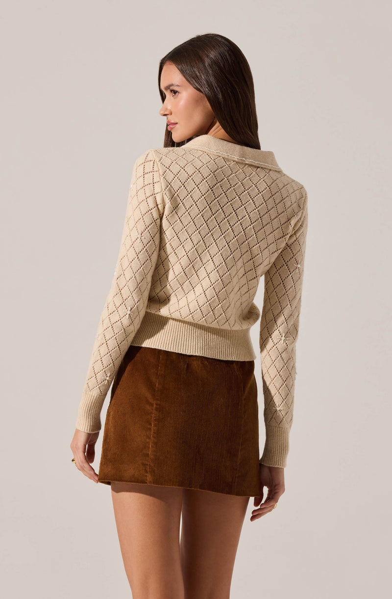 A woman is wearing a beige knit sweater with a diamond pattern and a ribbed hem and cuffs, paired with a brown corduroy skirt.