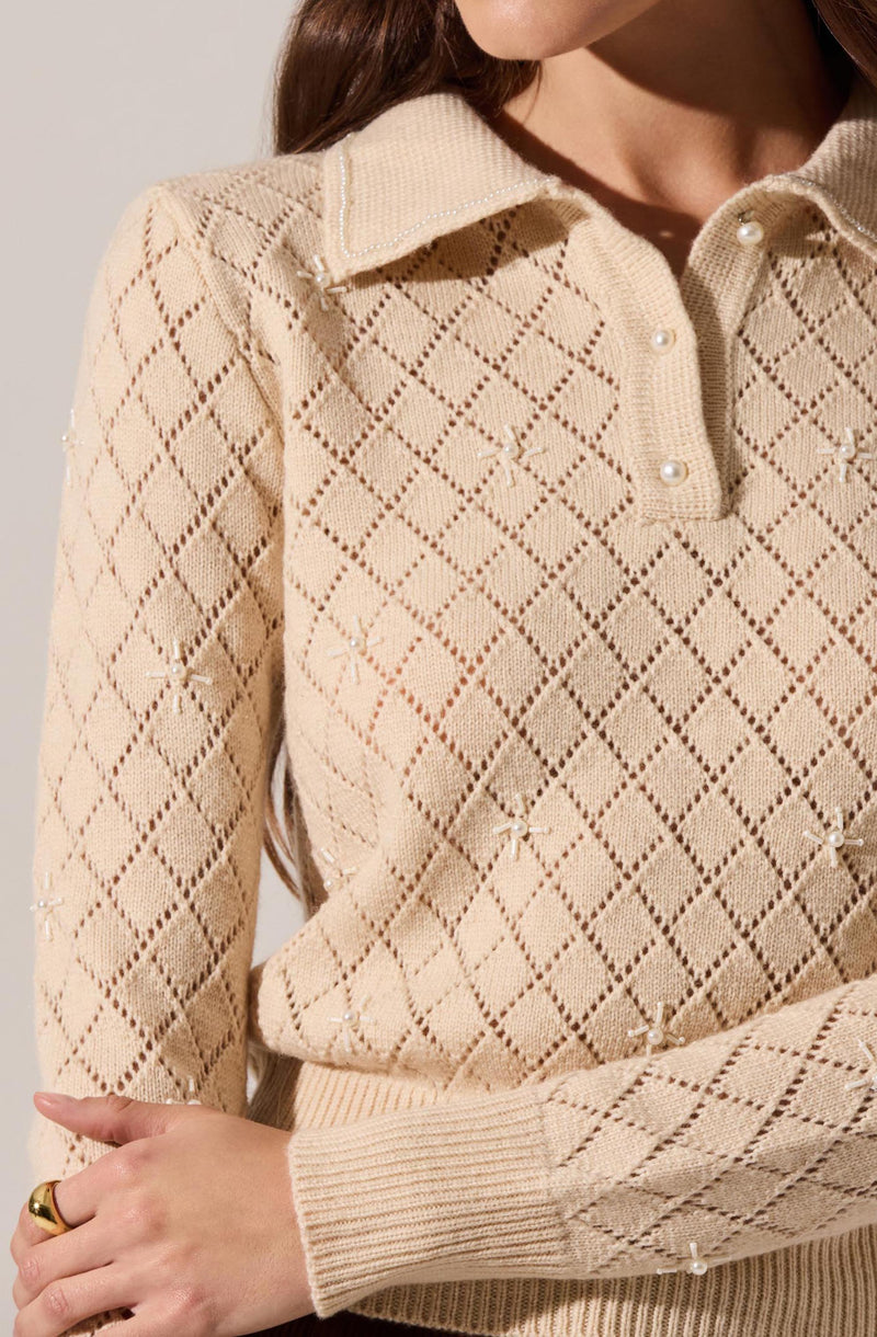 A woman is wearing a beige knitted sweater featuring a diamond pattern with small bows and pearl embellishments scattered throughout. The sweater has a collared neckline with a button-up front.