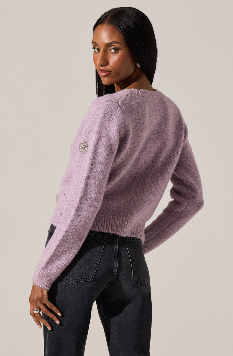A woman is wearing a soft, fuzzy, lavender-colored sweater with long sleeves. The sweater features a small decorative emblem on the left sleeve near the shoulder. She pairs it with high-waisted, black denim jeans.