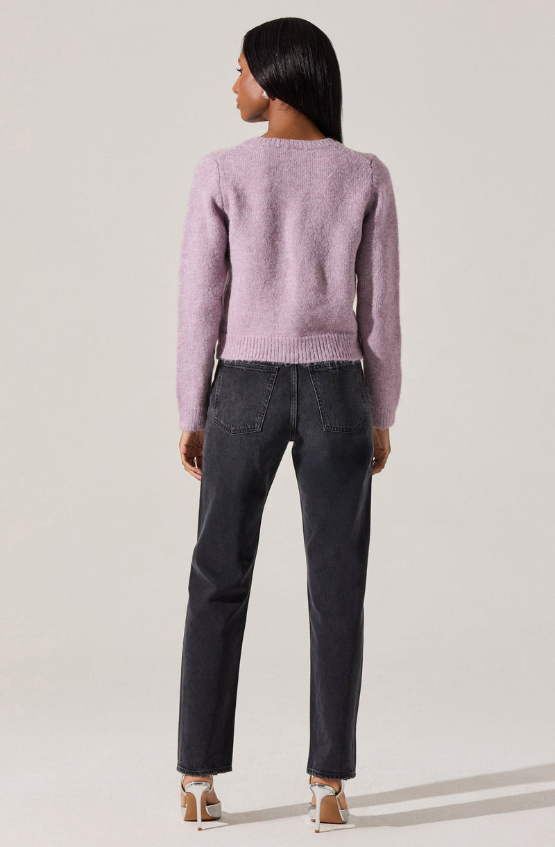 A woman is wearing a soft, fuzzy, light purple sweater with long sleeves and a round neckline, paired with high-waisted, slightly faded black jeans with visible stitching on the back pockets. She is also wearing nude-colored heels with a white ankle strap.