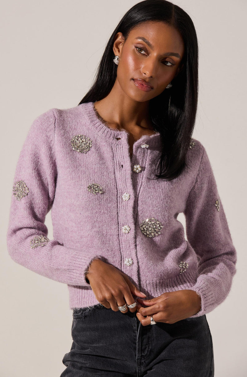 A woman is wearing a light purple, knit cardigan sweater adorned with clusters of rhinestones and floral embellishments. The cardigan has a round neckline and button-down front. She is also wearing dark-colored pants.