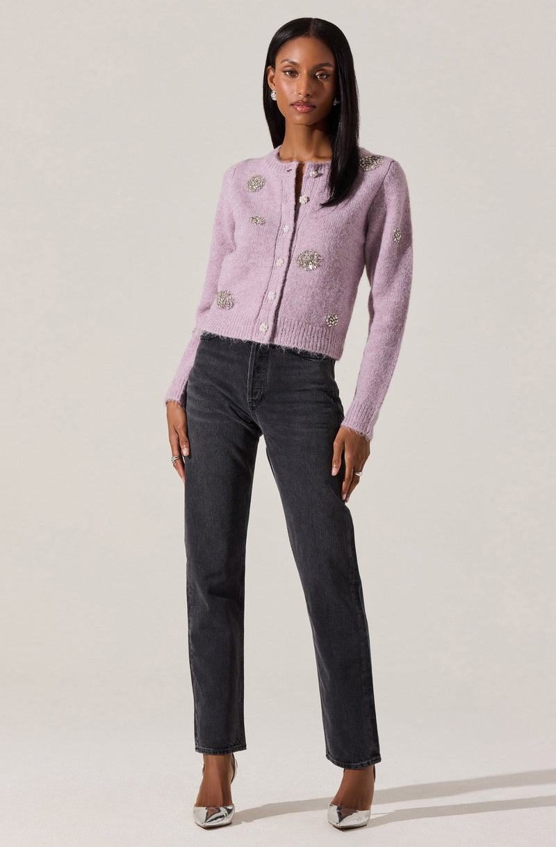 A woman is wearing a light purple, long-sleeved cardigan with decorative silver embellishments scattered across the front and sleeves. The cardigan has a round neckline and pearl buttons down the front. She is also wearing high-waisted, dark gray jeans and white pointed-toe heels with a pattern.