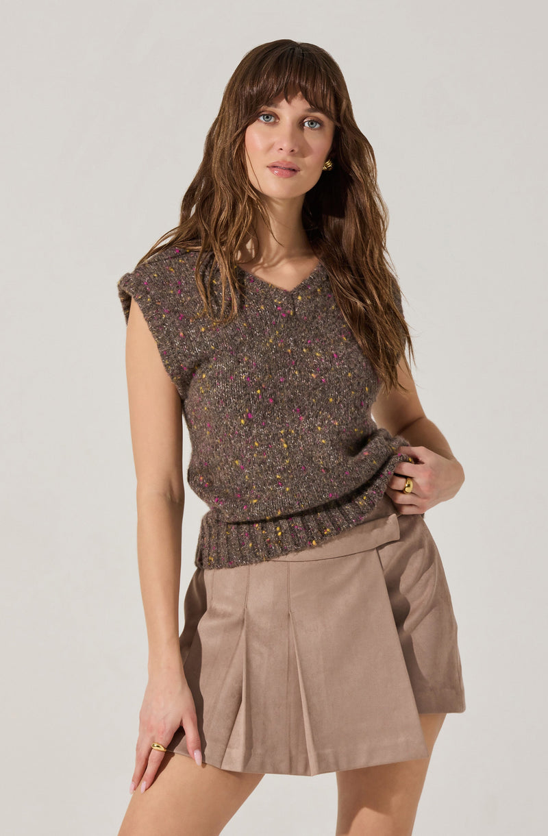 A woman is wearing a sleeveless, knitted sweater in a dark speckled pattern with pink, yellow, and green flecks. She pairs it with beige high-waisted shorts that have pleats in the front. She has wavy brown hair with bangs and is accessorized with gold earrings and rings.