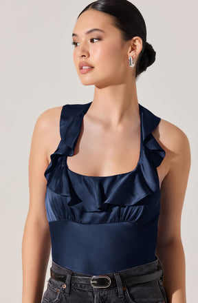 A woman is wearing a navy blue sleeveless top made of shiny satin fabric. The top features ruffled straps and a ruffled neckline. She is also wearing dark jeans with a black belt that has a metallic buckle. The woman has her hair styled in a low bun and is accessorized with large, silver hoop earrings.