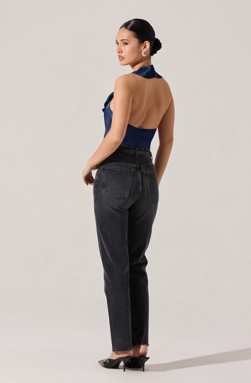 A woman is wearing a sleeveless, halter-style top in a shiny navy blue fabric. The top is backless and tied around her neck, exposing her back. She pairs it with high-waisted, dark-washed jeans and black high-heeled shoes.