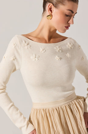 A woman is wearing a cream-colored knit sweater with textured floral appliqués featuring three-dimensional flowers around the neckline and shoulders. The sweater has a wide, rounded neckline and long sleeves. She pairs it with a beige, pleated midi skirt made of lightweight fabric. She has gold, leaf-shaped earrings and her hair is styled in a sleek bun.