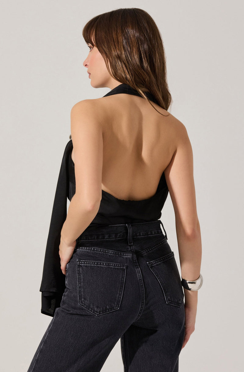 A woman is wearing a sleeveless, backless black top and high-waisted black jeans. She has a silver wristwatch on her left wrist and is holding a black jacket over her left arm.